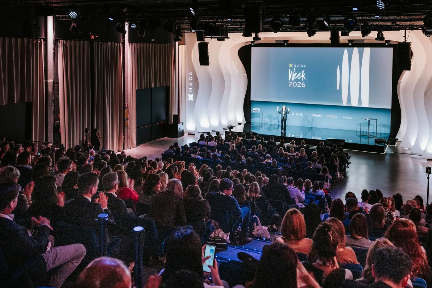 Packed auditorium at NAOS Week 2026 in Brussels, a presenter on stage in front of curved white panels and a 'NAOS Week 2026 Pulse' screen, cinematic stage lighting.