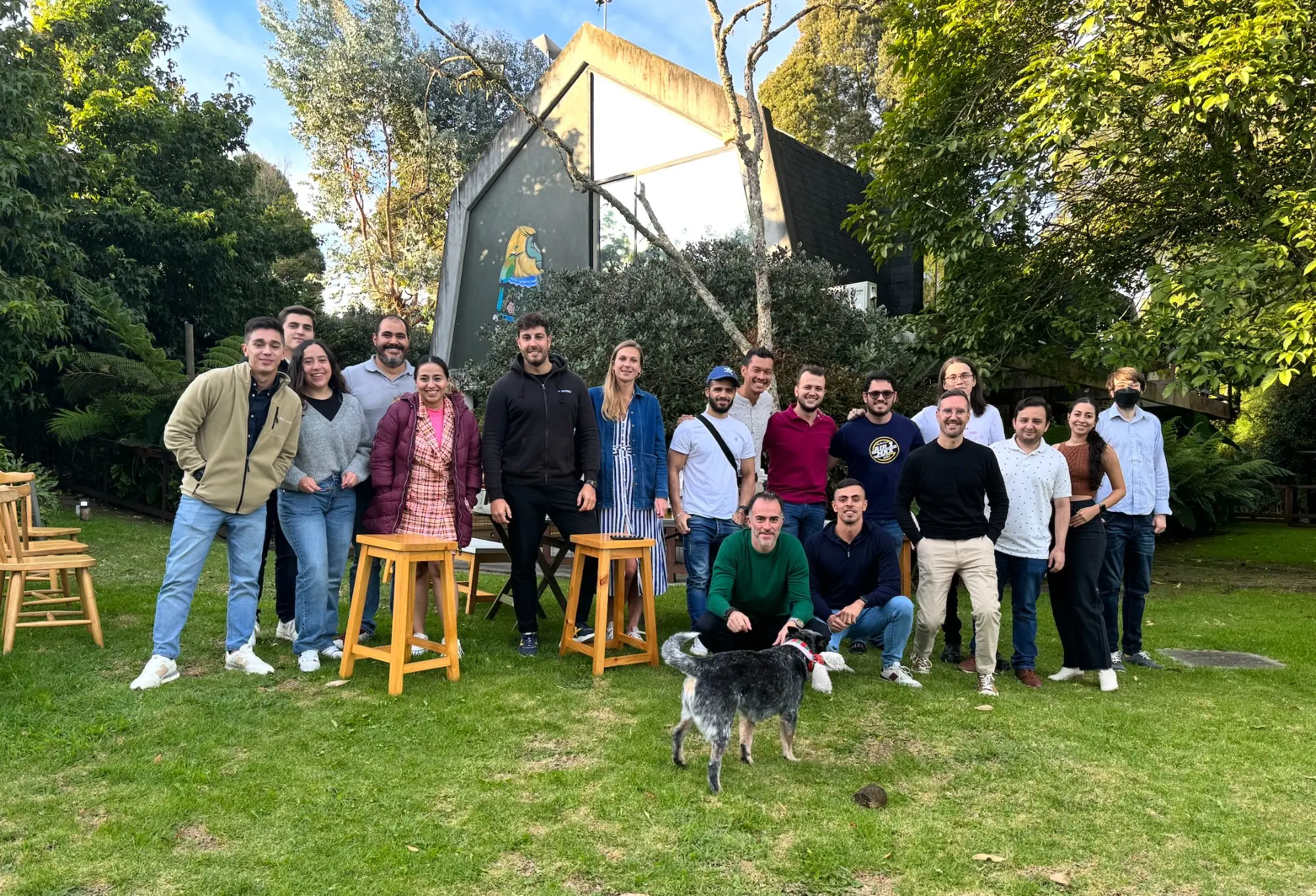 The Eventtia team gathered together outdoors at a remote team gathering, around stools on a grass lawn with a dog in front.
